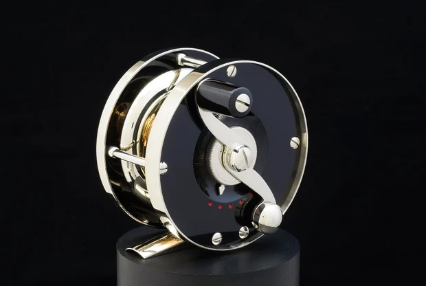 A Wagner small-batch fly reel: nickel silver frame with a black anodised side plate, knurled drag knob and a curved counterbalanced handle, photographed against black.